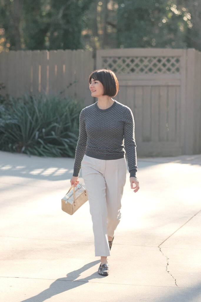 Kelly walking in charcoal polka dot fitted sweater with cream ankle pants, plaid loafers, and gold handbag — full-length walking shot of in-between season petite outfit