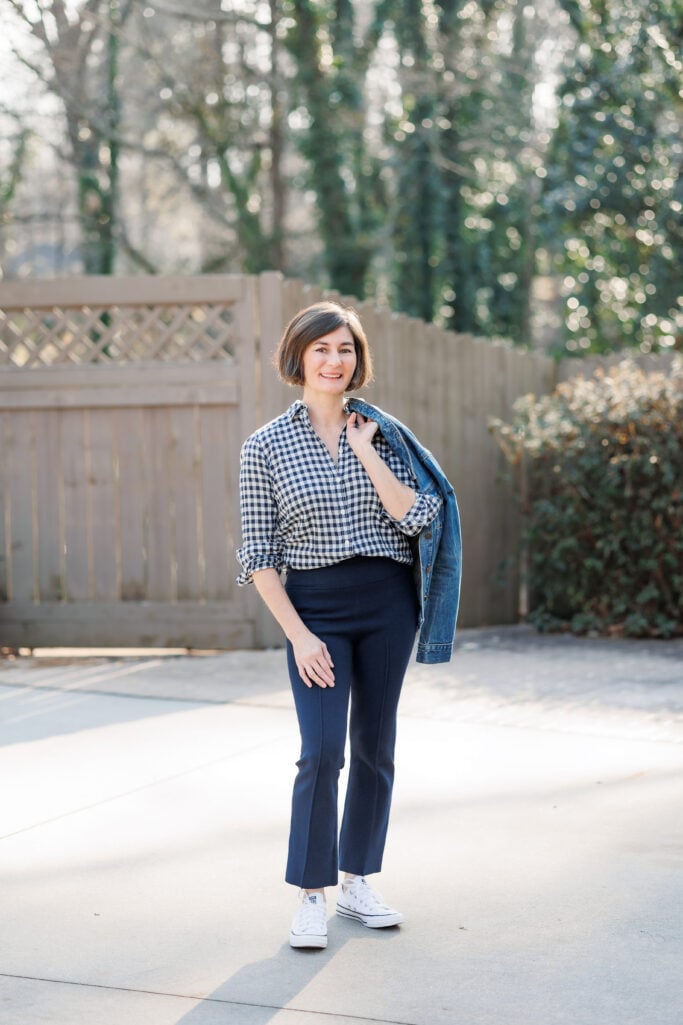 Kelly styling a navy gingham button-up with navy kick flare pants and white Converse sneakers, denim jacket over shoulder