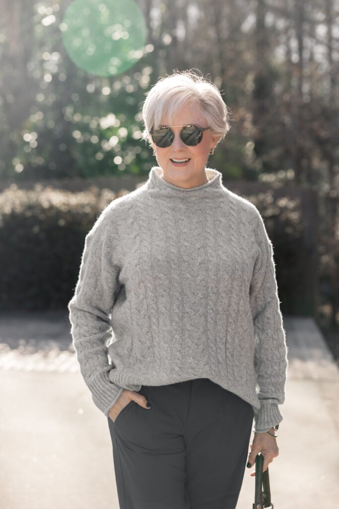 Woman over 50 wearing light gray cable knit mock neck sweater with charcoal tailored trousers and aviator sunglasses