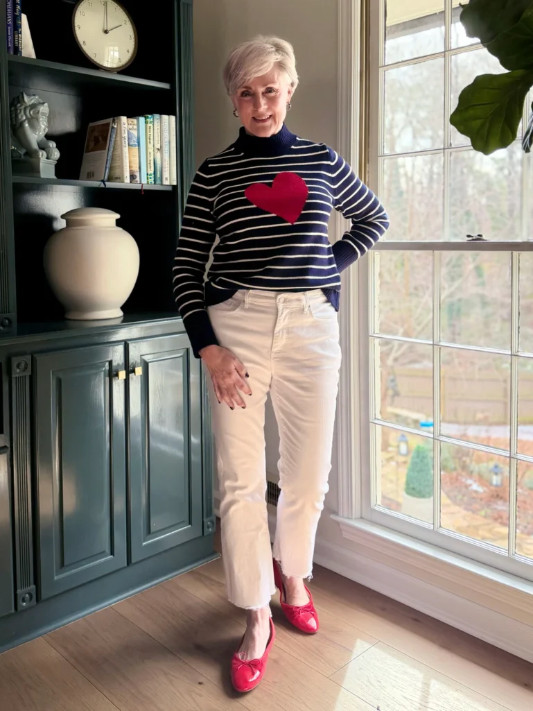 Woman over 50 wearing a navy and white horizontal striped mockneck sweater with red heart detail, cream denim pants, and red patent ballet flats, showing how to wear horizontal stripes stylishly over 50.