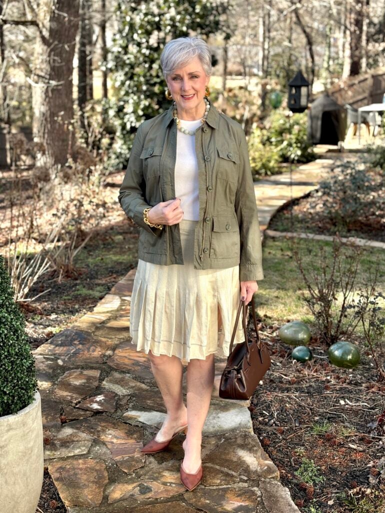 Beth Djalali wearing a green utility jacket over a white top with a pleated cream skirt, gold and pearl necklace, brown leather heels, and a brown leather bag