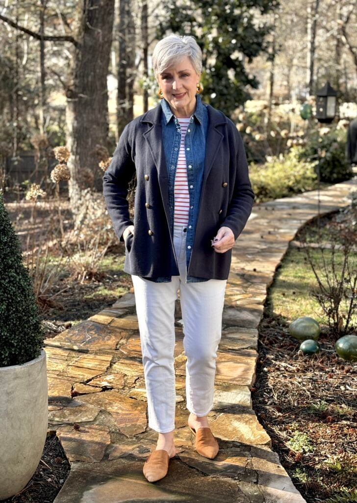 Beth Djalali wearing a navy sweater blazer with gold buttons layered over a denim shirt and red striped tee, styled with white jeans and tan suede mules