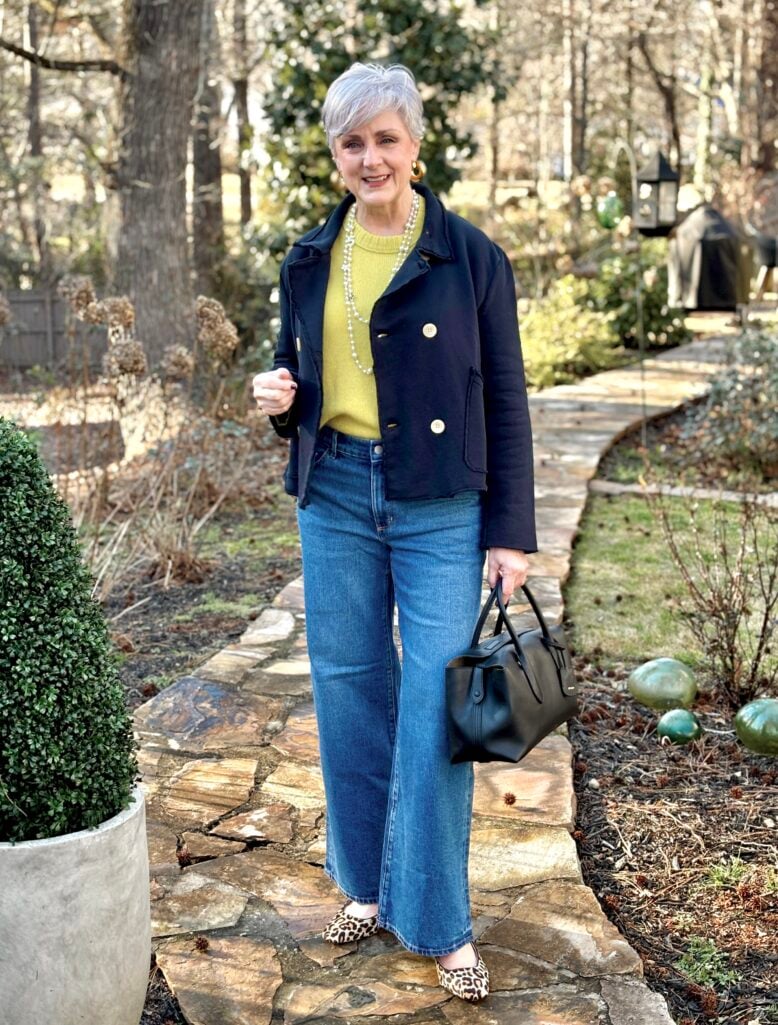 Beth Djalali wearing a navy cropped peacoat with gold buttons over a chartreuse sweater, styled with wide-leg medium-wash jeans, leopard print flats, a long pearl necklace, and a black leather bag