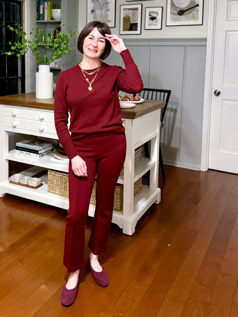 Kelly wearing a burgundy cashmere crewneck sweater, burgundy flare-leg pants, layered gold chain necklaces with a heart pendant, and plum suede ballet flats, hand brushing her hair by the kitchen island