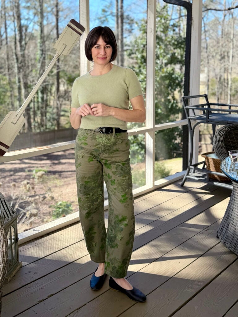 Kelly wearing a sage green cashmere shrunken tee, high-rise green floral print barrel jeans, a black covered buckle leather belt, and navy Day Glove ballet flats on the screened porch