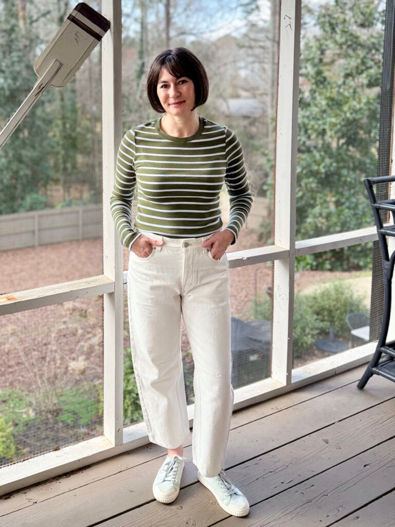 Kelly wearing an olive-and-white striped ribbed long-sleeve tee, cream barrel-leg jeans, and mint mesh sneakers, hands in pockets on the screened porch