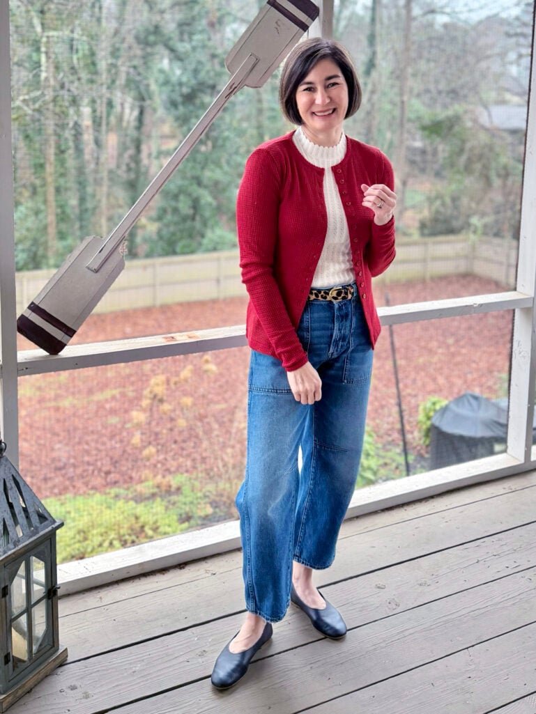Kelly wearing a red cardigan over a cream pointelle top, barrel leg jeans with a leopard print belt, and navy ballet flats on the screened porch