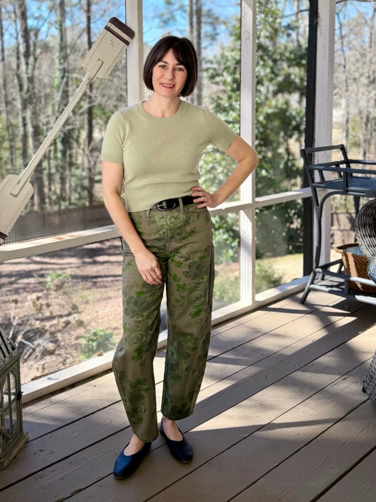 Full-length view of Kelly with hand on hip in her green cashmere tee and floral barrel jeans, sunshine streaming across the porch