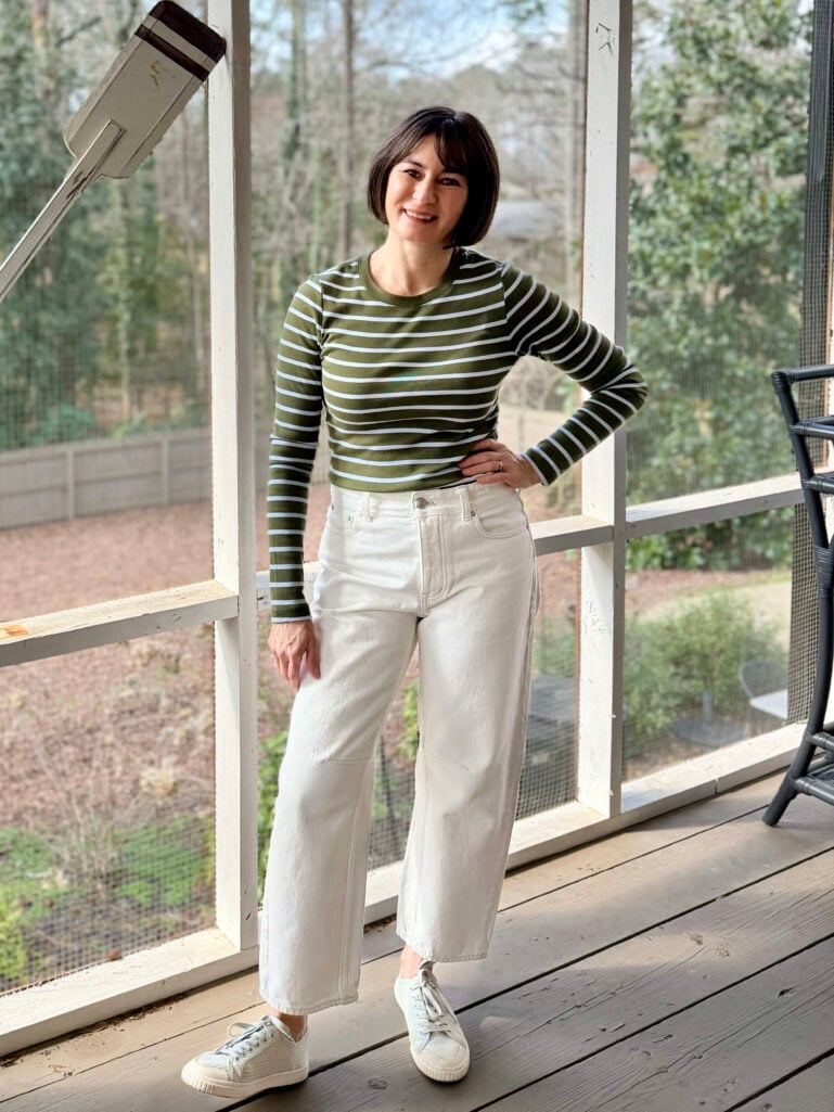 Full-length view of Kelly in her olive striped tee and cream barrel-leg jeans, hand on hip, smiling on the screened porch with winter greenery visible through the screens
