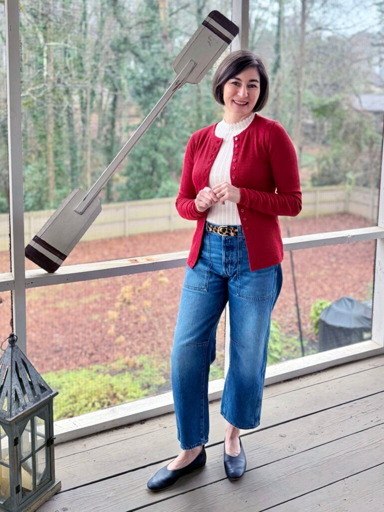 Full-length view of Kelly in her red cardigan and jeans, hands together, smiling on the screened porch