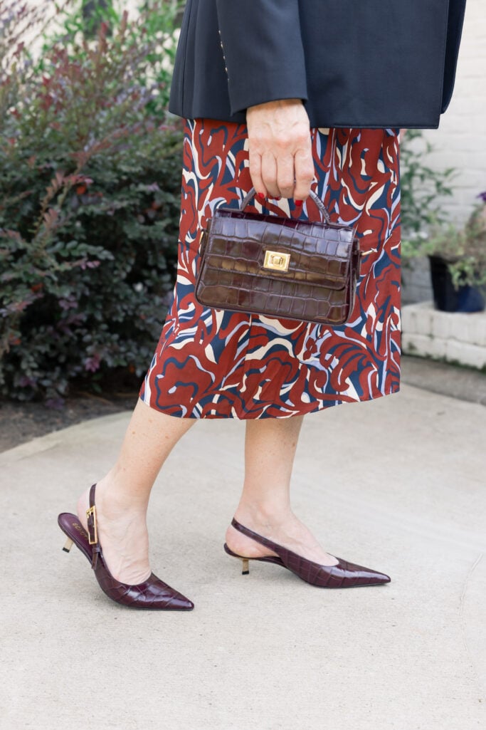 Close-up of burgundy croc-embossed pointed-toe slingback kitten heels with gold buckle detail, paired with navy blazer, rust and navy printed midi skirt, and brown croc-embossed top-handle bag