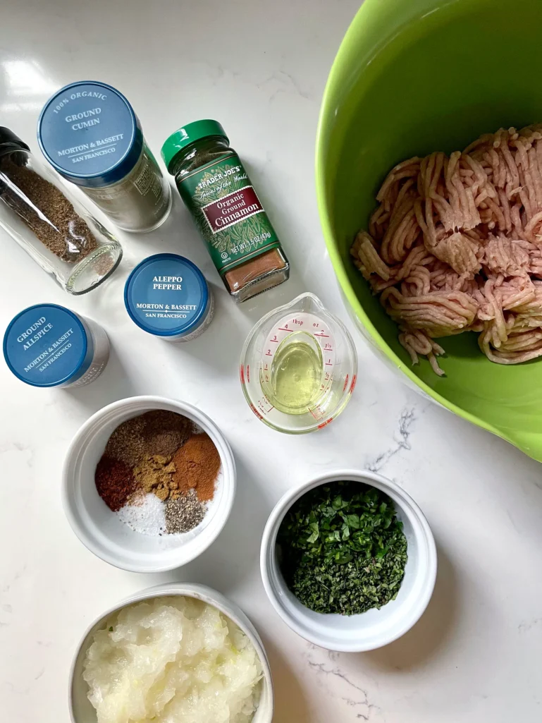 Chicken kofta ingredients laid out: ground chicken in a green bowl, spice jars, grated onion, chopped herbs, measured oil, and mixed spices in small bowls