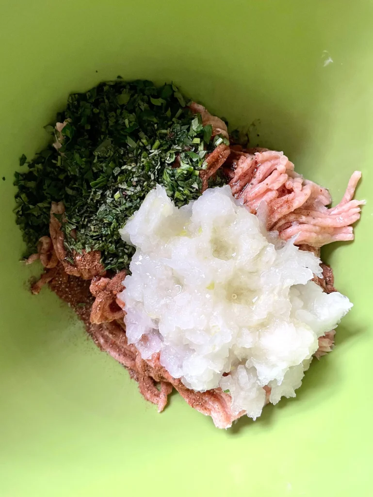 Ground chicken with grated onion, chopped herbs, and spices before mixing in a green bowl