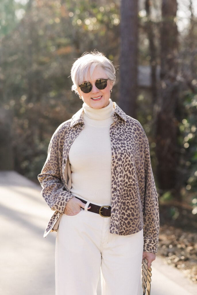 Woman over 50 wearing leopard print shirt jacket open over ivory turtleneck sweater with cream wide-leg jeans, dark leather belt, and tortoiseshell sunglasses