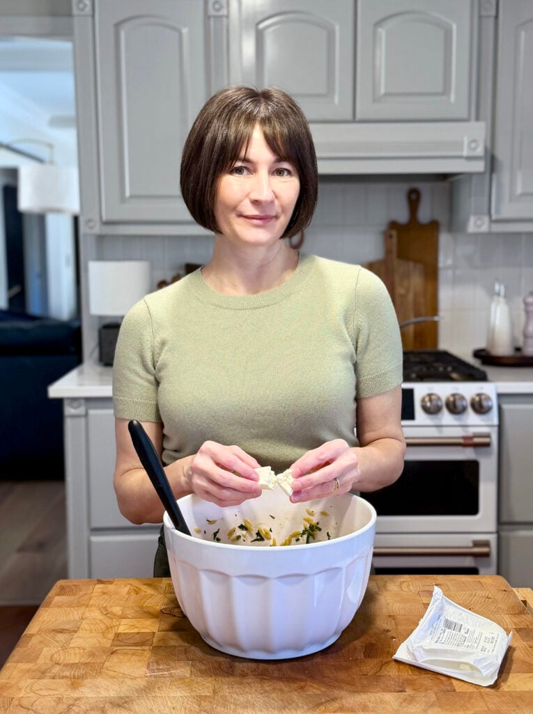 Kelly crumbling feta cheese into a white mixing bowl of Lemon Herb Orzo in the kitchen, wearing her sage green cashmere tee