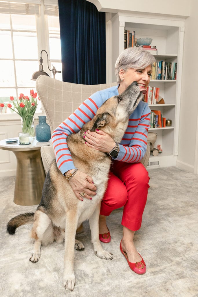 Archive photo of Beth and Oscar from a 2022 Valentine's Day Fridays with Oscar post, Beth in a blue-and-red striped sweater and red pants laughing as Oscar gives her a kiss, red tulips and bookshelves in the background