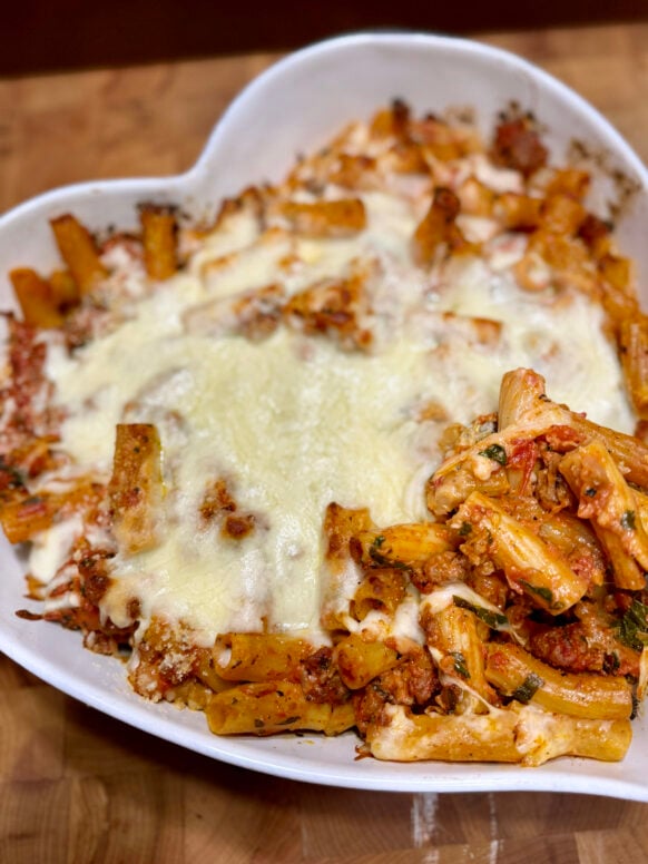 Rigatoni pasta bake with melted mozzarella, Italian sausage, and vodka cream sauce in a white heart-shaped baking dish, golden and bubbling from the oven