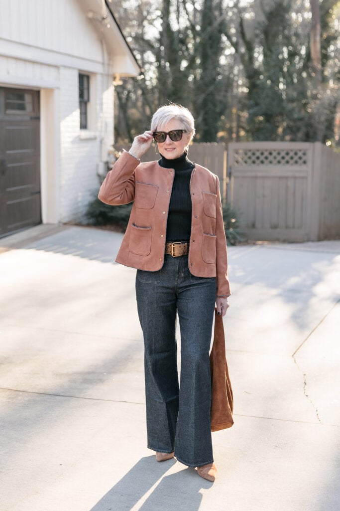 Woman over 50 with silver hair wearing cognac suede jacket with stitch detail over black turtleneck, dark wash high-rise wide-leg jeans with cognac leather belt, tan suede pointed-toe flats, brown suede hobo bag, tortoiseshell sunglasses, and gold hoop earrings
