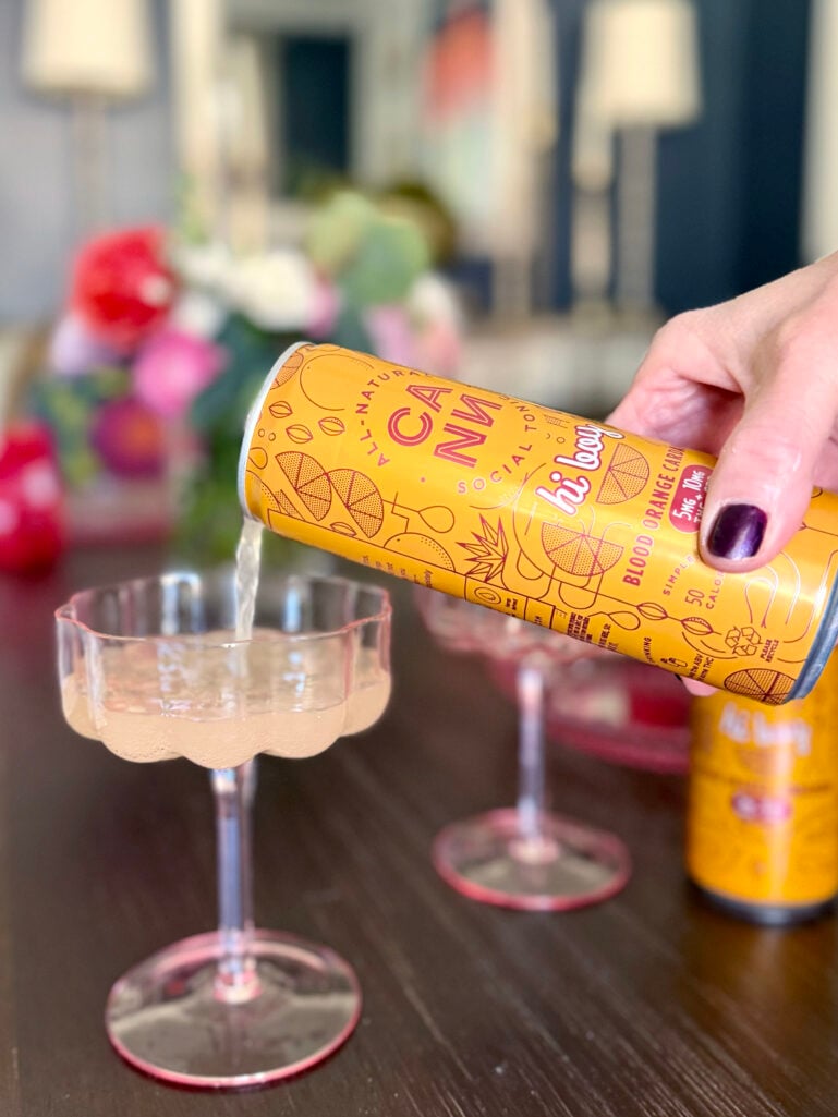 Beth's hand pouring a Cann Blood Orange Cardamom social tonic into a pink scalloped coupe glass, dark nail polish visible, with Valentine's flowers blurred in the background
