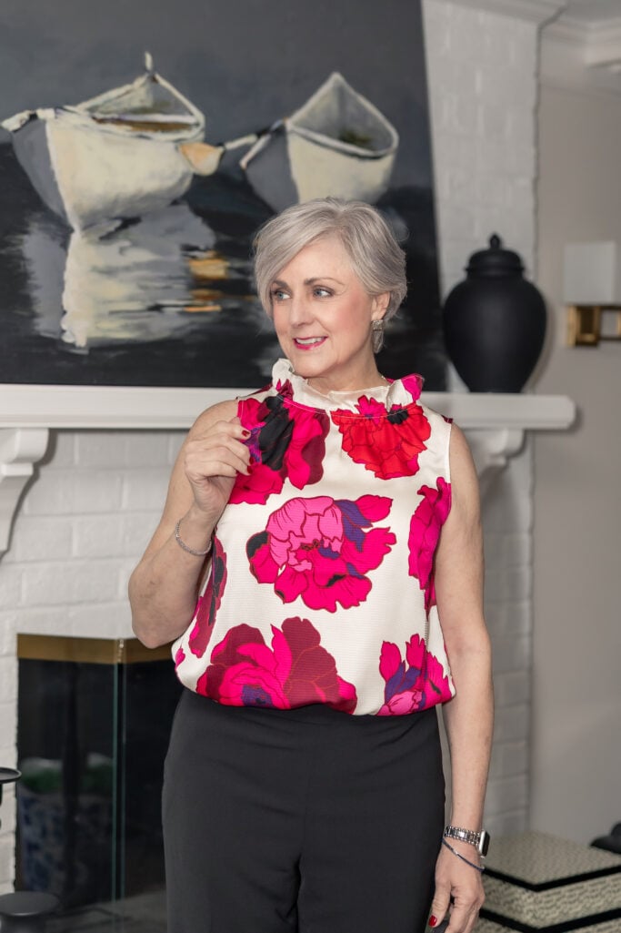 Beth in a pink and red floral sleeveless blouse with a ruffle neckline and black pants — incorporating red into a print for women over 50