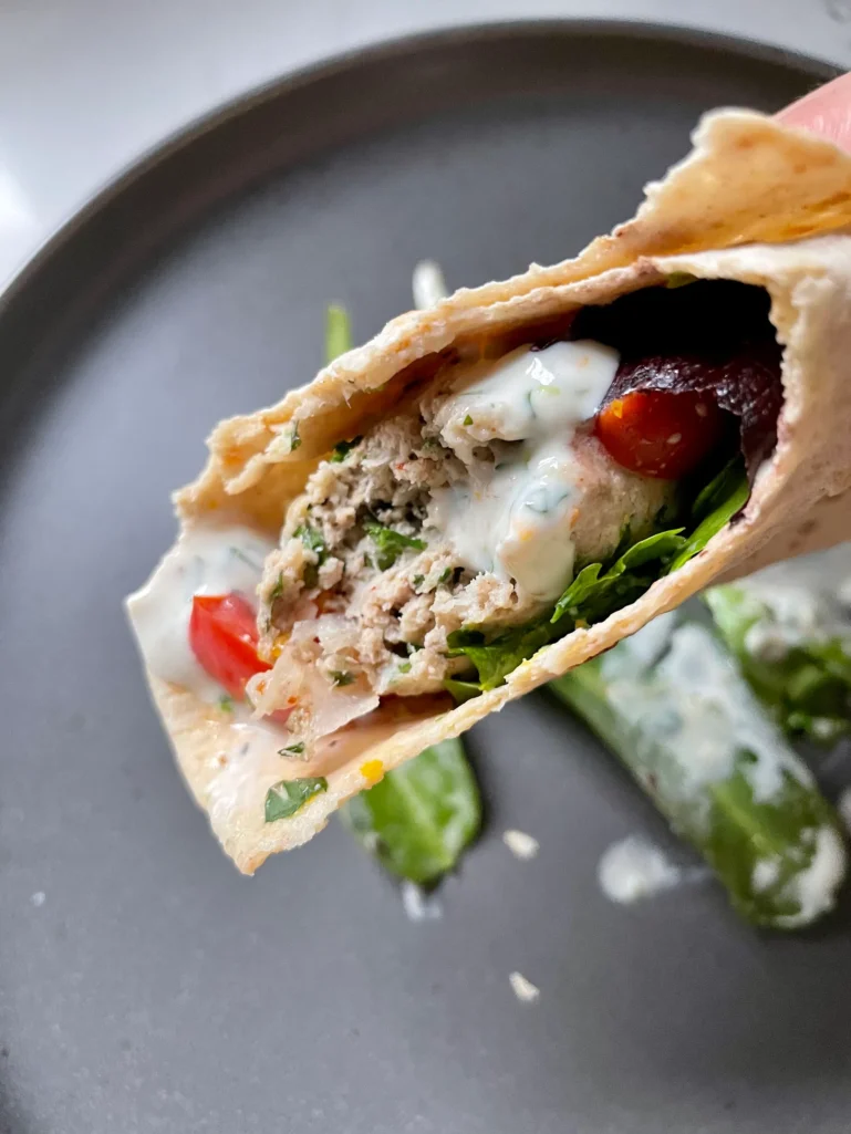 Hand holding a rolled lavash wrap filled with chicken kofta, yogurt sauce, greens, and grape tomatoes
