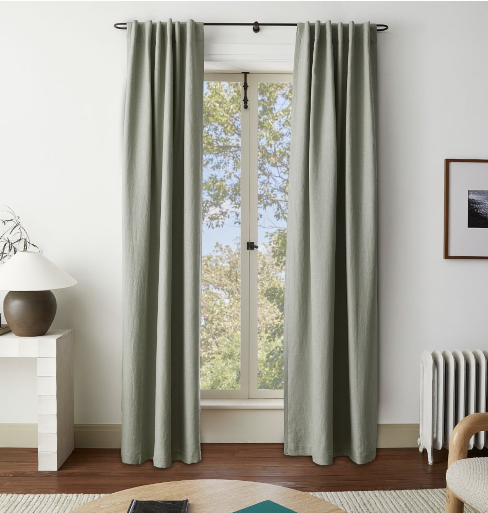 Sage green Belgian flax linen blackout curtains hanging from a black curtain rod, softly filtering daylight in a modern living space.