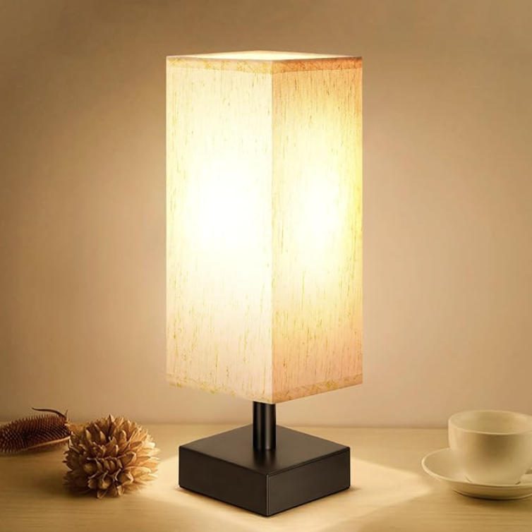 Modern rectangular bedside table lamp with a soft fabric shade glowing warm yellow light, placed on a wooden nightstand next to neutral decor.