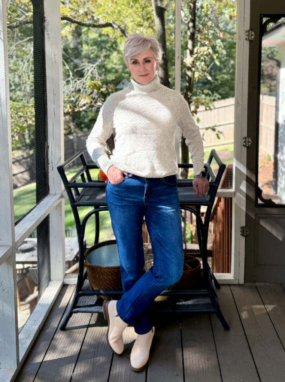 Woman with short silver hair wearing a cream speckled turtleneck sweater, dark blue straight-leg jeans, and beige ankle boots standing on a screened porch with greenery in the background.