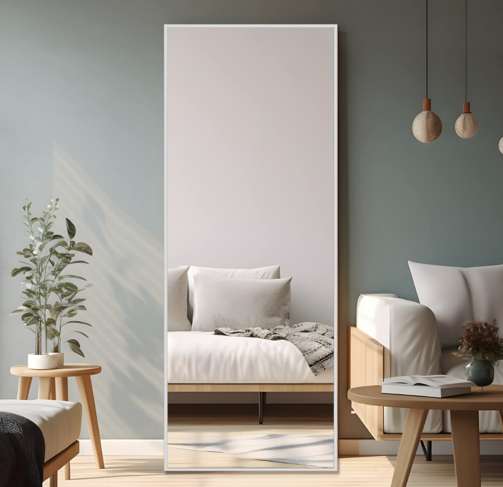 Tall rectangular full-length floor mirror with slim white frame, reflecting a minimalist bedroom with neutral bedding, wood accents, and soft natural light.