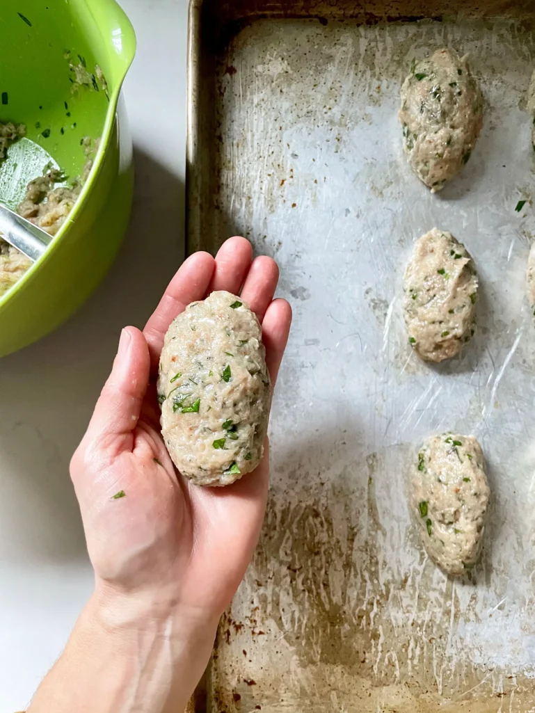 Hand holding an oval-shaped raw chicken kofta over a sheet pan with more shaped kofta ready to broil