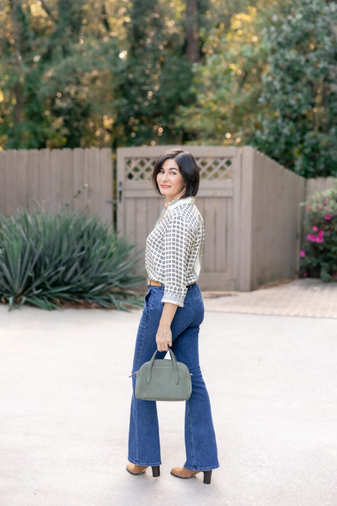 Woman with brown bob haircut wearing cream and navy geometric print blouse tucked into dark wash flare jeans, tan suede ankle boots, sage green structured handbag