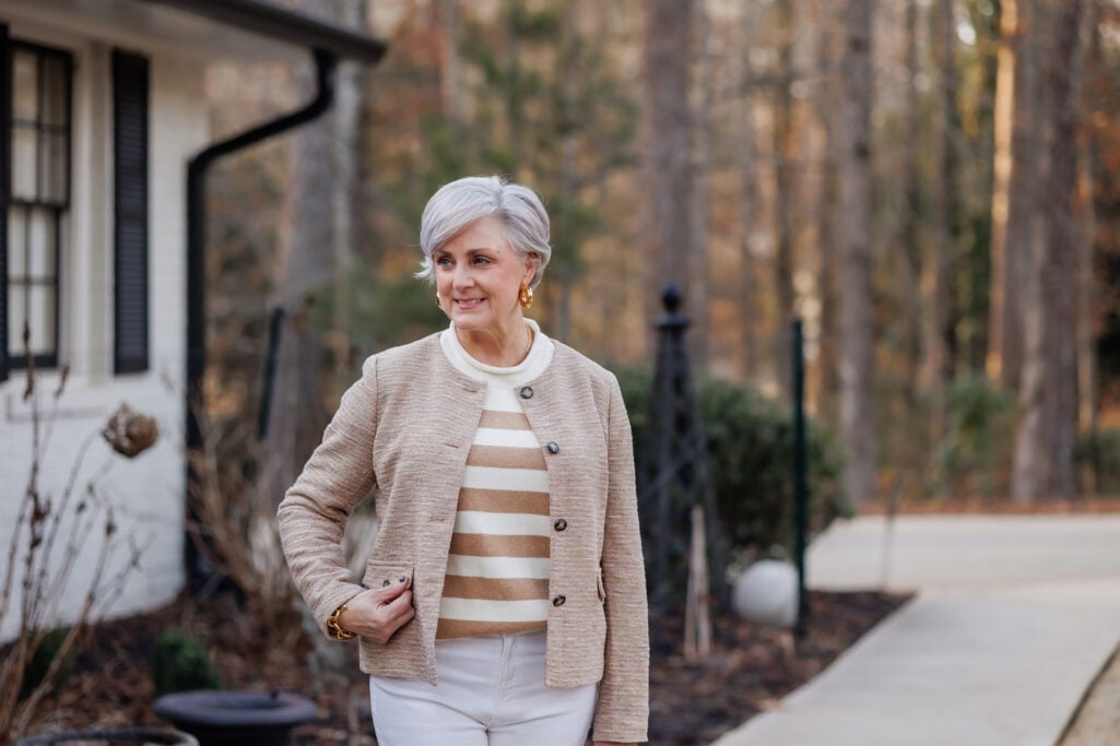 Woman over 50 wearing camel and ivory striped sweater layered under oatmeal boucle knit jacket with gold hoop earrings and gold link bracelet