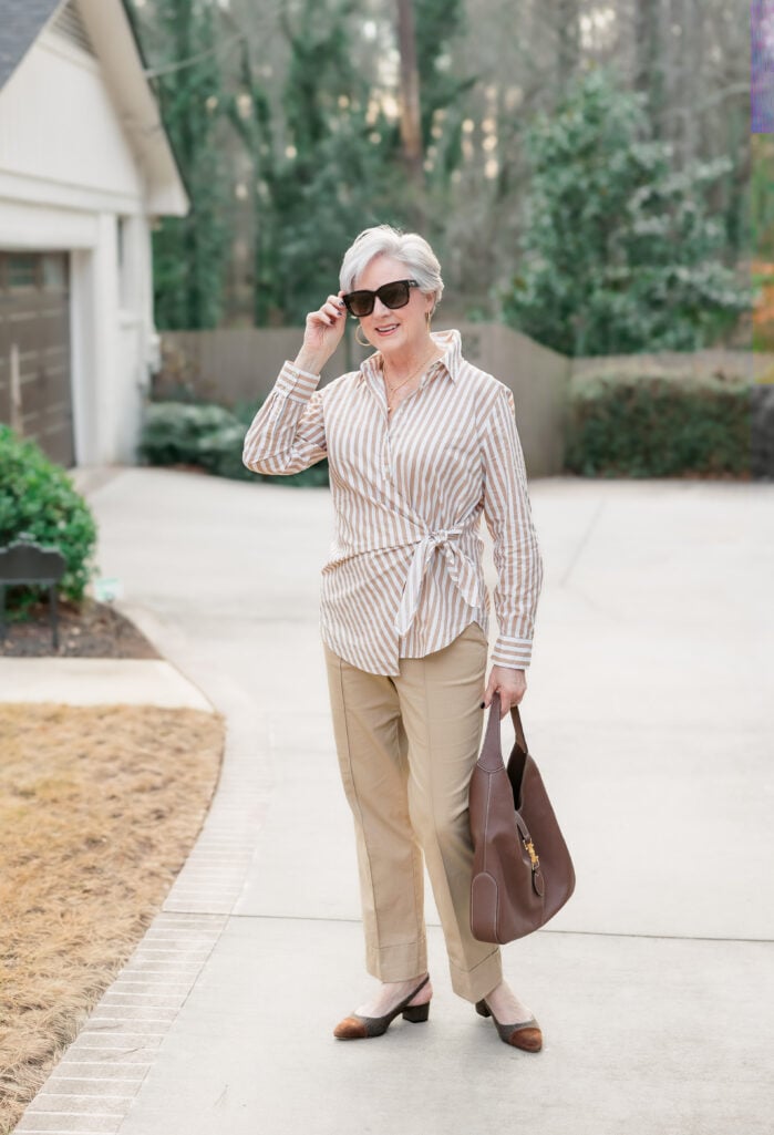 Woman over 50 styling vertical stripes in a tan and white tie-front poplin blouse with khaki ankle pants, chocolate brown leather hobo bag, tweed slingback heels, oversized sunglasses, and gold hoop earrings for a chic spring outfit.