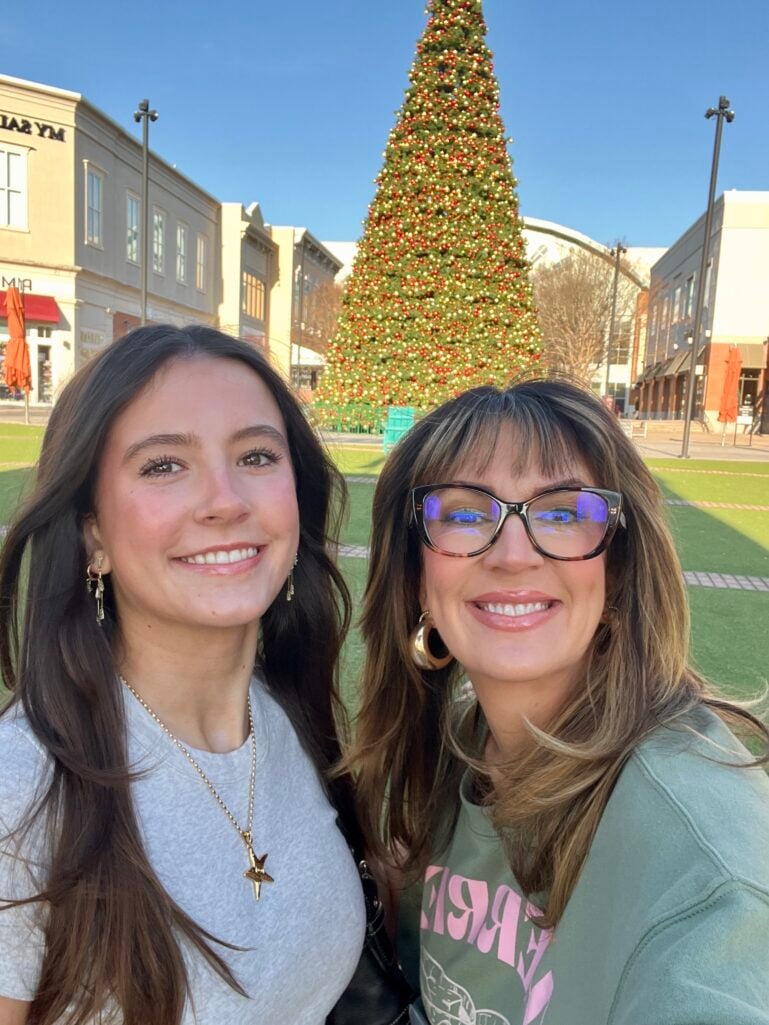 Mother and teenage daughter Vivian smiling together in outdoor shopping center selfie with large decorated Christmas tree behind them, daughter wearing gray t-shirt with gold star pendant necklace and small gold hoop earrings, mother wearing sage green graphic sweatshirt, tortoise cat-eye glasses, gold hoop earrings, both with natural glowing makeup and big smiles