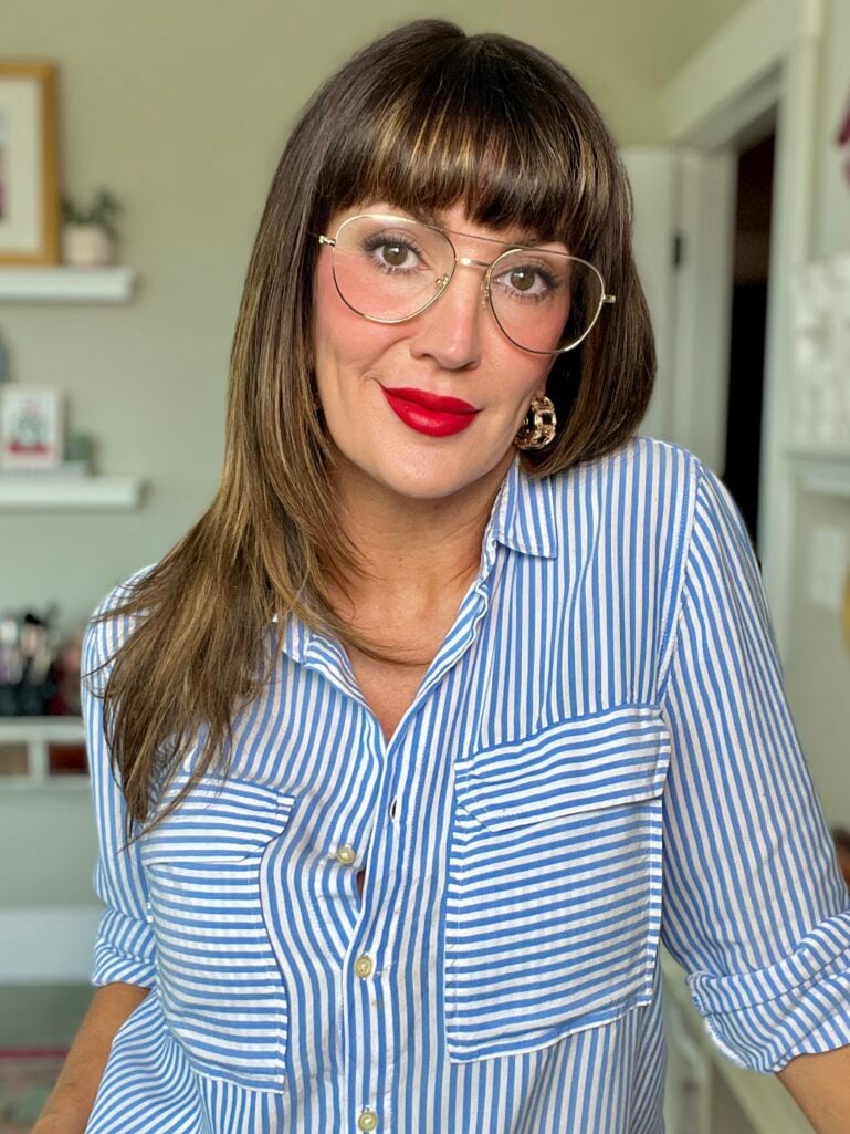 Woman wearing bold matte red lipstick with blue and white striped button-down shirt, gold aviator glasses, gold chunky chain link hoop earrings, soft rosy cheeks, sleek straightened hair with bangs