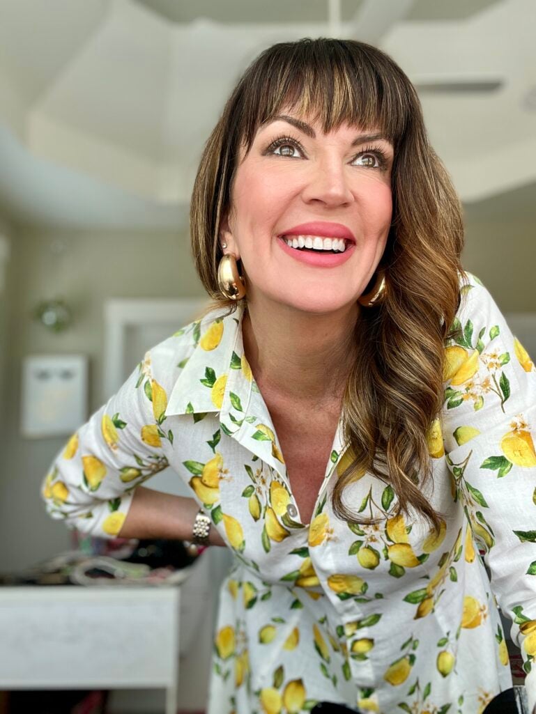 Woman laughing with big smile looking upward, wearing white linen button-down shirt with yellow lemon print, gold chunky hoop earrings, coral-pink lipstick, pearl bracelet, demonstrating that a smile is the best makeup accessory
