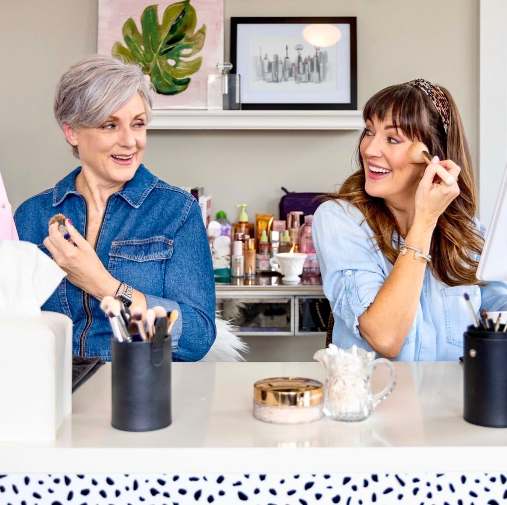 Two women doing makeup together at vanity, woman with silver pixie cut wearing denim zip-up shirt holding makeup brush and lipstick, woman with brown highlighted hair and bangs wearing chambray shirt and leopard print headband applying powder with brush, both smiling at each other, makeup products and brushes on desk, monstera leaf art and cityscape print on wall behind