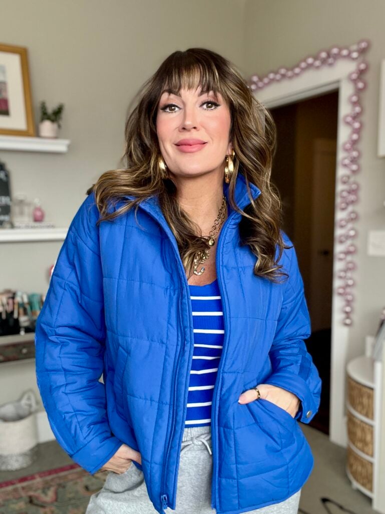 Woman with highlighted brown hair and curtain bangs wearing royal blue quilted puffer jacket over blue and white striped sweater, hands in jacket pockets with gray joggers, layered gold chain necklace with charms and gold hoop earrings, standing in front of pink beaded mirror