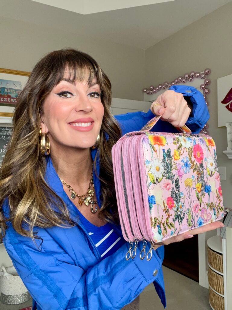 Woman in royal blue puffer jacket holding pink floral-patterned zippered pencil case with handle, case has four zipper compartments, gold hoop earrings and layered charm necklace visible, smiling at camera