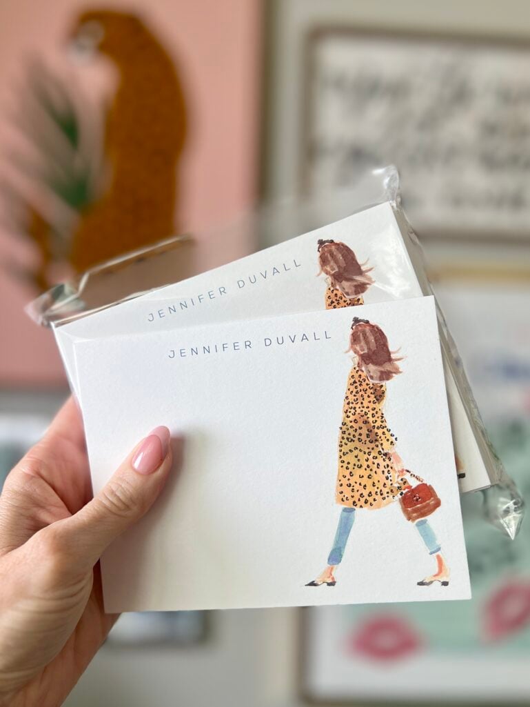 Hand holding two sets of personalized stationery cards featuring watercolor illustration of a woman in leopard print coat carrying a handbag, cards read Jennifer Duvall, pink nails visible, colorful wall art in blurred background