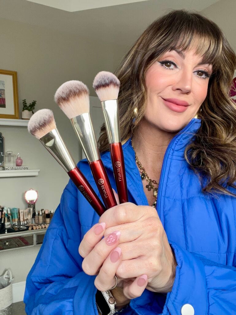 Woman in royal blue puffer jacket holding three BK Beauty face brushes with dark red handles and silver ferrules, brushes fanned in hand showing different head shapes, soft pink press-on nails with flower accent nail, beauty products visible on shelf in background