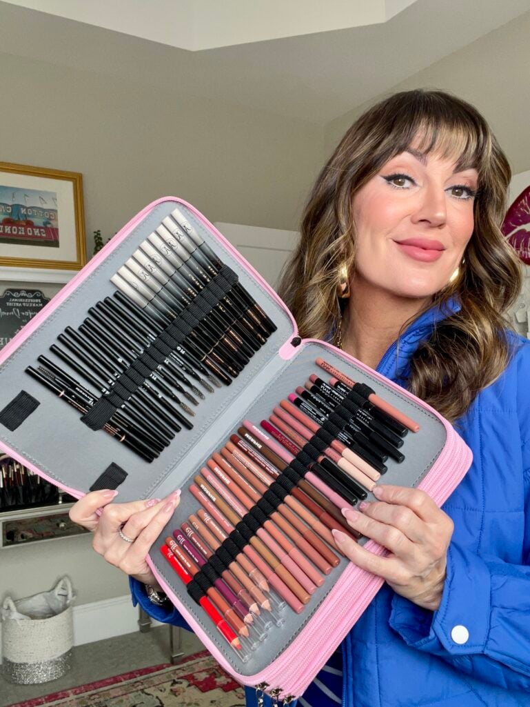 Woman in royal blue puffer jacket holding open pink floral 200-slot pencil organizer repurposed for makeup, left side filled with black eyeliner and eyebrow pencils, right side organized with lip liners in nude and pink shades, multiple elastic loops visible