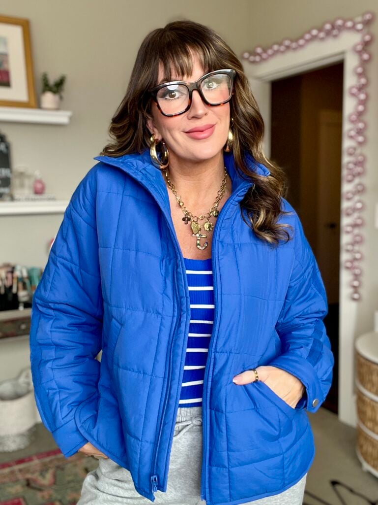 Woman wearing royal blue puffer jacket with tortoiseshell Peepers glasses, blue and white striped top, gray joggers, hand in jacket pocket, gold charm necklace and hoop earrings, standing in front of beaded pink mirror with beauty products on shelf