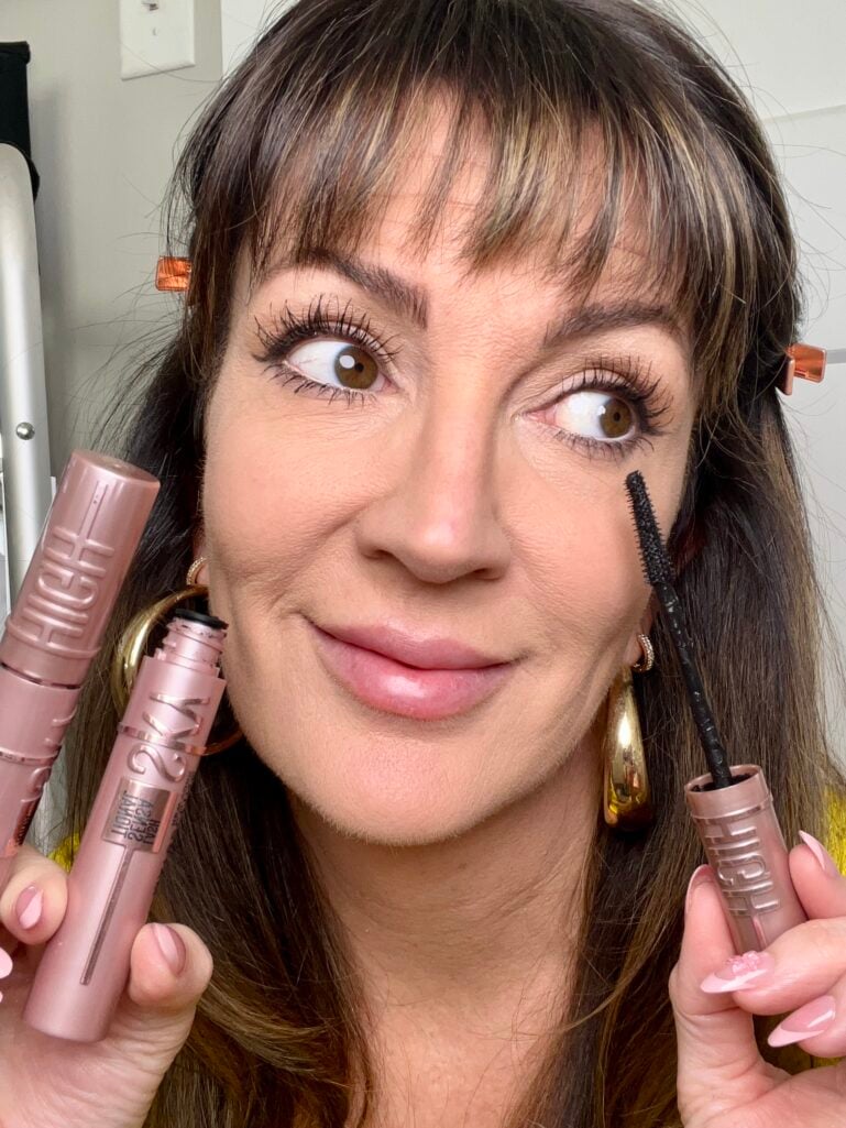 Woman holding Maybelline Lash Sensational Sky High mascara tube and showing flex tower wand brush near face, demonstrating dramatically lengthened lashes, wearing yellow sweater, gold puffy hoop earrings, copper hair clips, pink French tip nails, looking to the side to show lash transformation
