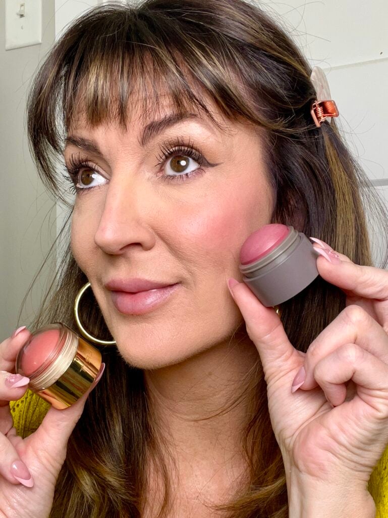 Woman holding Merit Flush Balm in gold and gray twist packaging near flushed cheeks showing applied rosy glow, wearing yellow sweater, gold puffy hoop earrings, copper hair clips, pink French tip nails, luminous skin with dewy flush visible