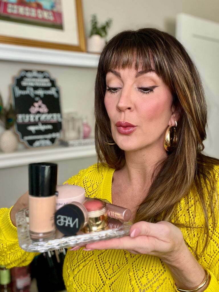 Woman looking down at wavy glass mirror tray holding iconic makeup products including Nars concealer, Armani foundation, Laura Mercier powder, Merit blush, and Clinique lipstick, wearing bright yellow pointelle knit sweater, gold puffy hoop earrings, rosy cheeks, mauve-pink lips, chalkboard sign and shelving visible in background