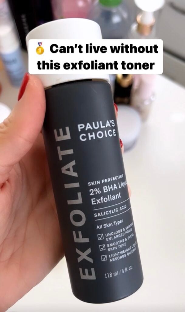 Hand holding Paula's Choice Skin Perfecting 2% BHA Liquid Exfoliant bottle in dark gray packaging with white cap, text overlay reading "Can't live without this exfoliant toner" with medal emoji, red manicure visible, vanity products blurred in background