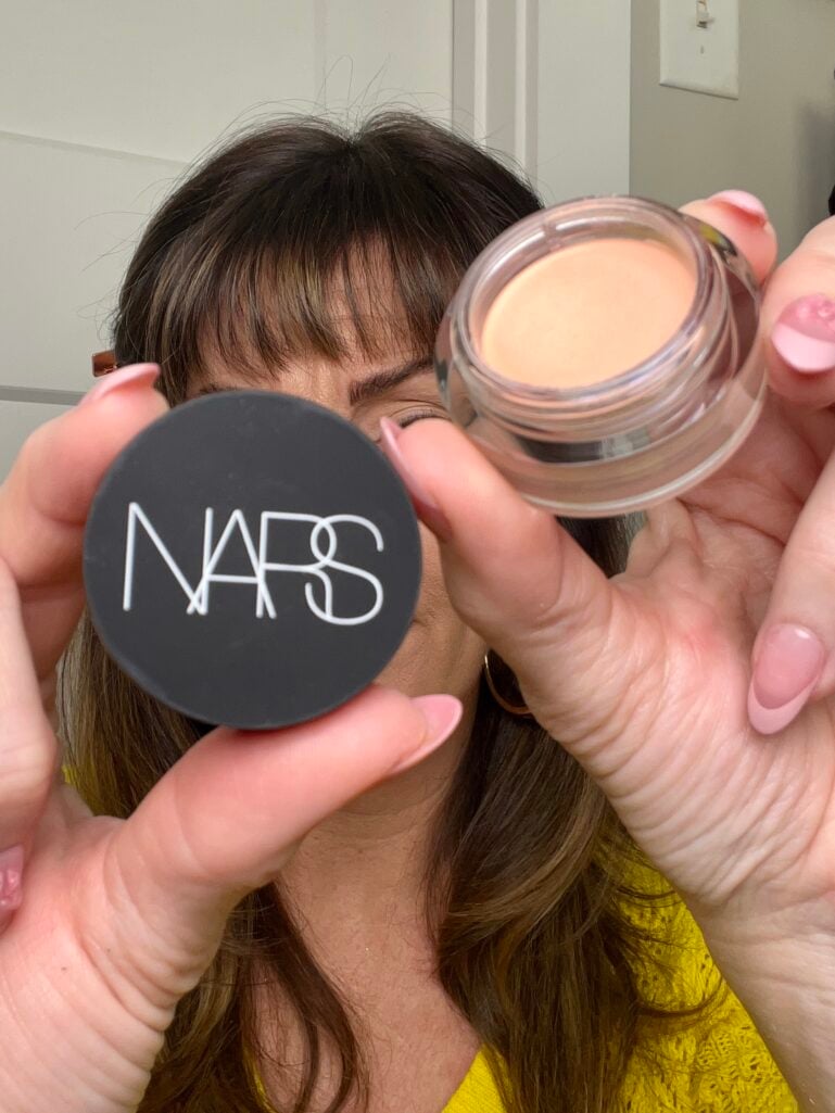 Woman holding Nars Soft Matte Complete Concealer pot with black lid in one hand and open jar showing light peachy shade in other hand, covering face, wearing yellow sweater, pink French tip press-on nails with heart detail, copper hair clips holding back bangs
