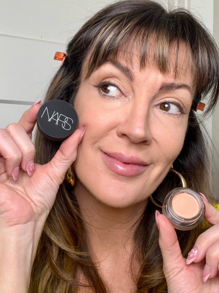 Woman holding Nars concealer lid near cheek and open pot showing shade Creme Brulee, wearing yellow sweater, gold puffy hoop earrings, copper hair clips, pink French tip nails, natural skin with concealer applied, looking to the side