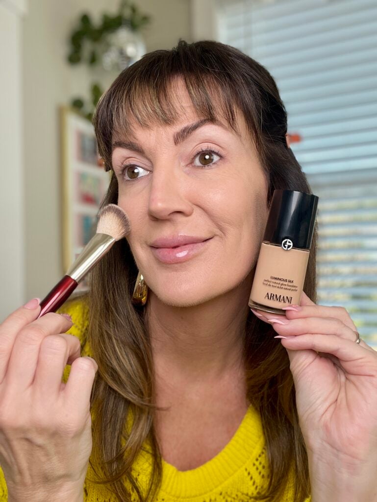 Woman applying foundation with BK Beauty burgundy-handled brush while holding Armani Luminous Silk Foundation bottle in shade 4.5, wearing yellow sweater, gold puffy hoop earrings, pink French tip press-on nails, natural skin mid-application, trailing plant and gallery wall visible in background
